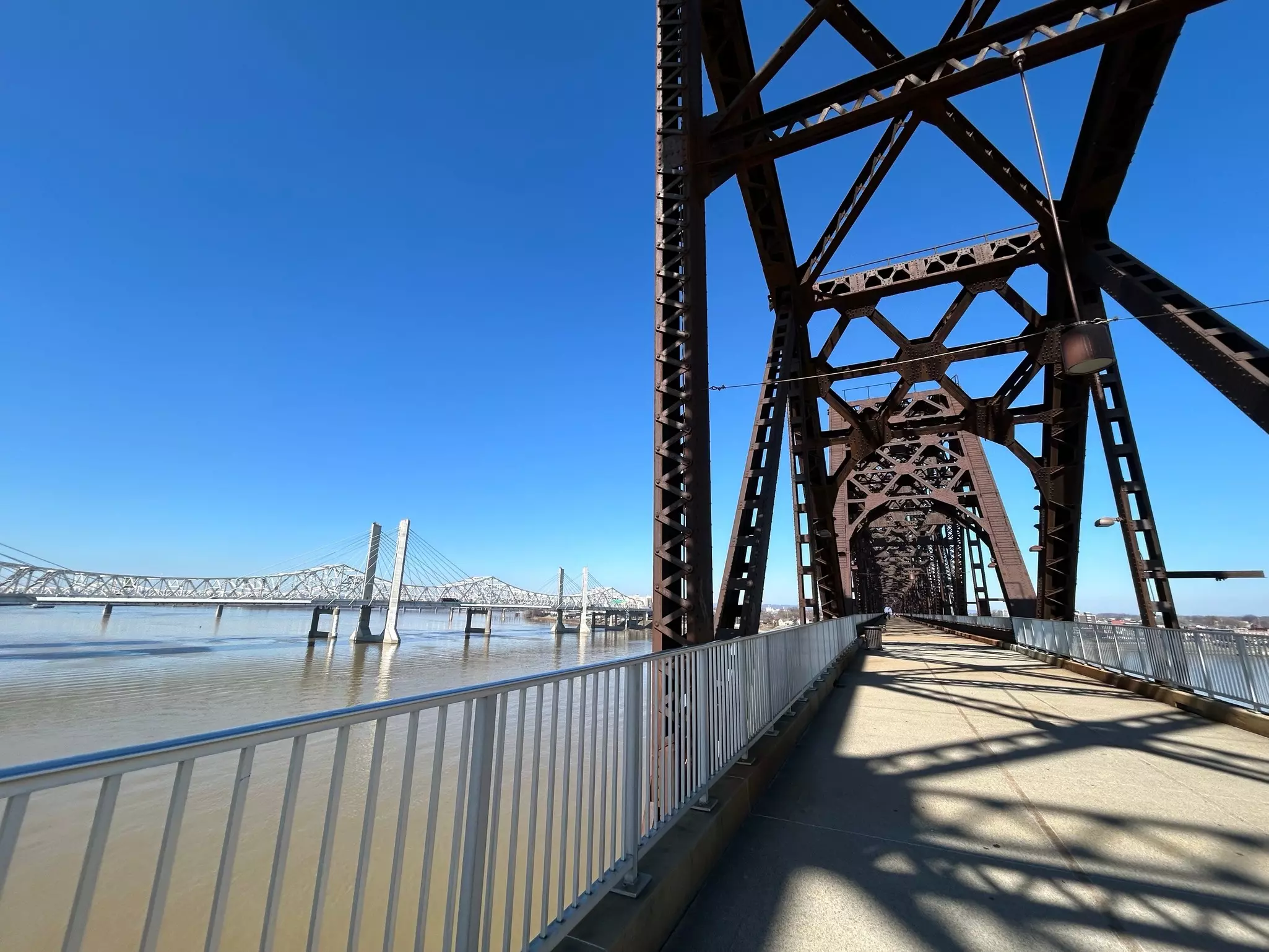 Metal-trussed bridge with cement walkway and larger bridge in the distance on a sunny day.