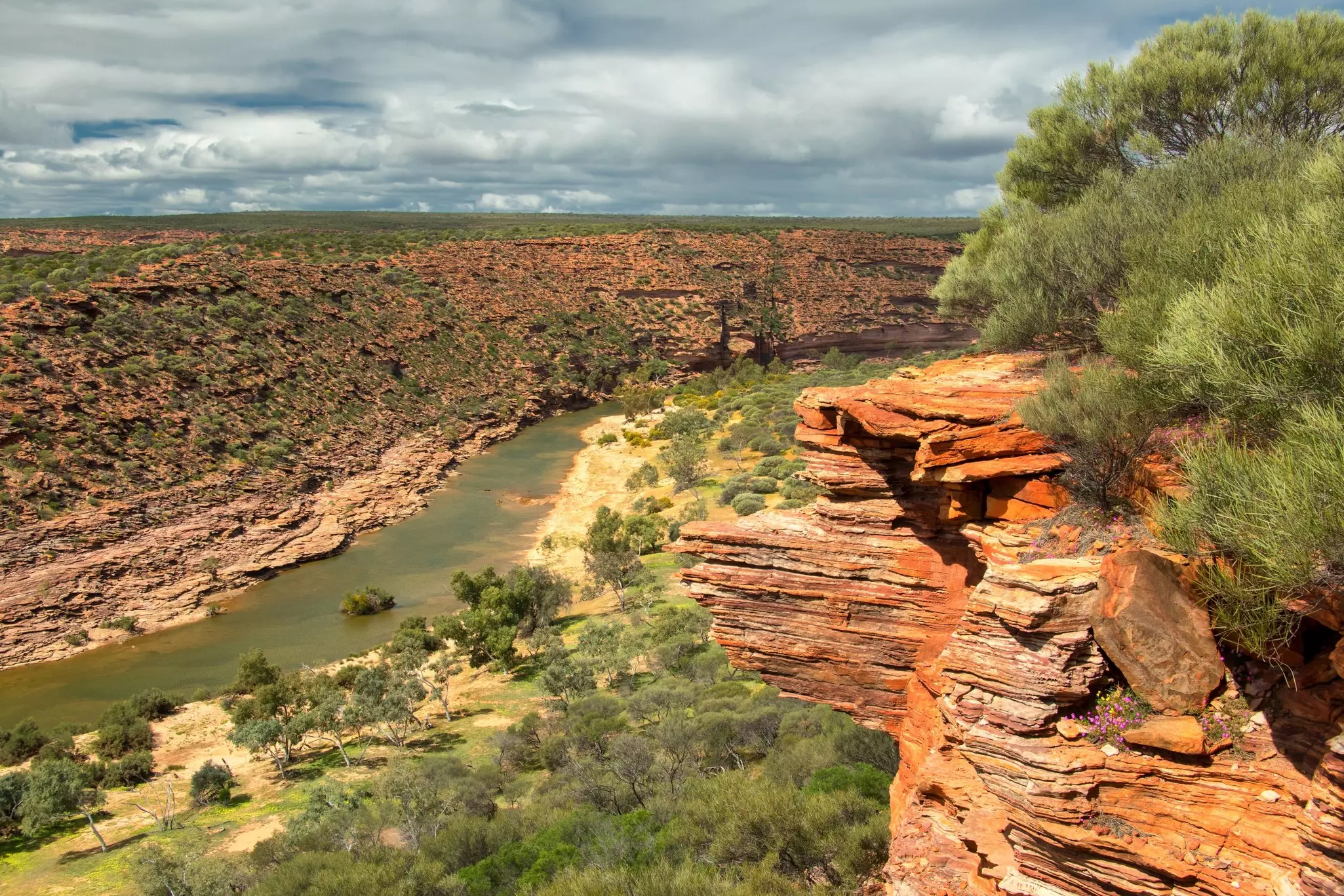 Green water cuts through a red rock cliff on one side and rolling hills covered in green scrub on the other