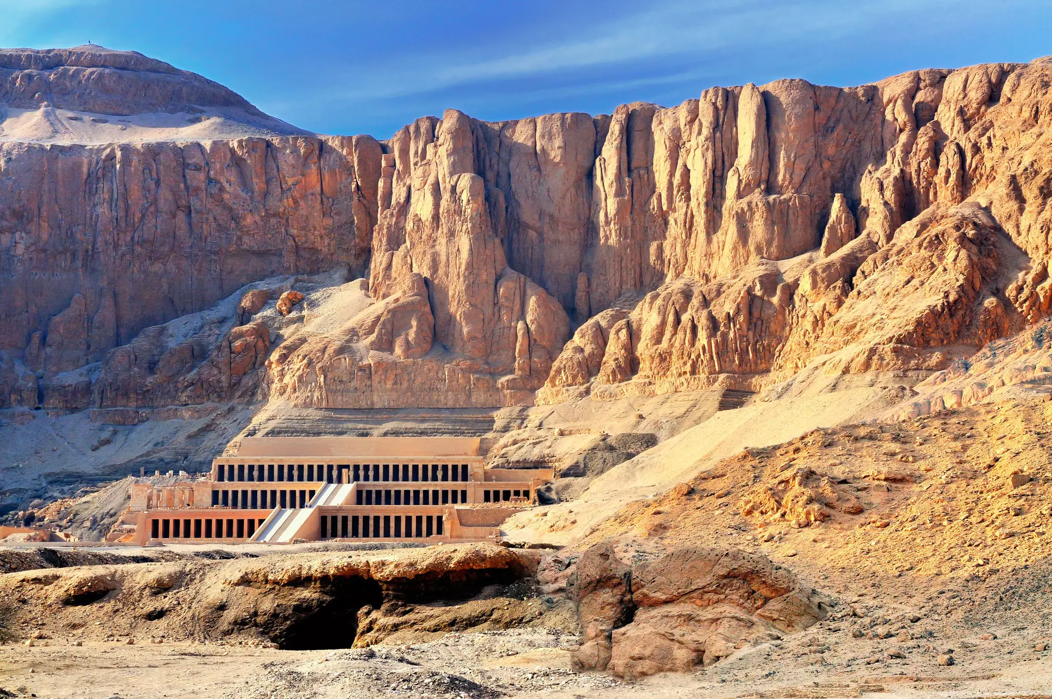 A tiered rectangular temple built into the base of a sandstone cliff in desert.