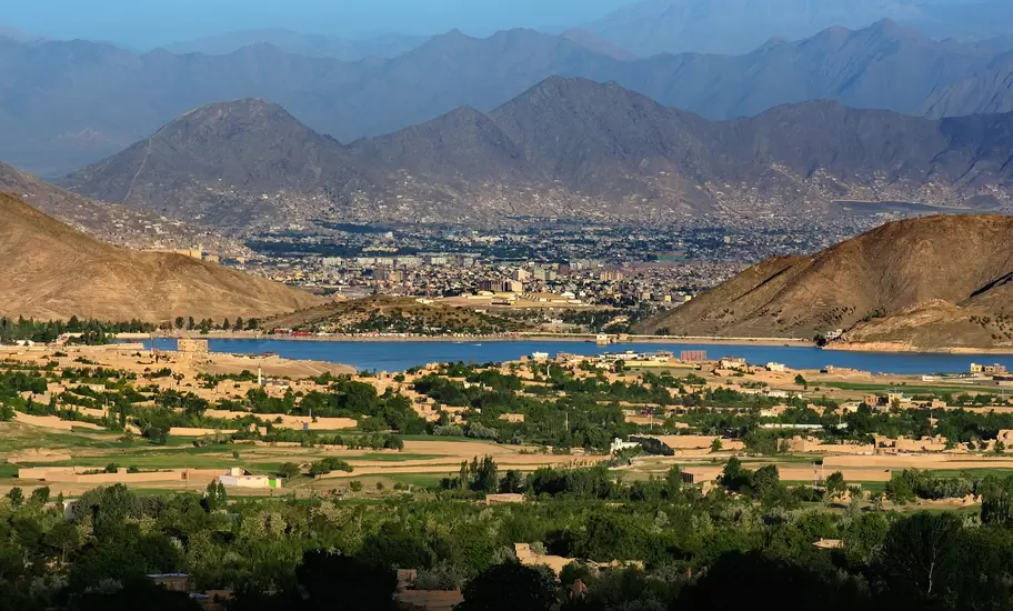 Kabul city sprawling below arid mountains, seen across a blue reservoir and green orchards, Afghanistan