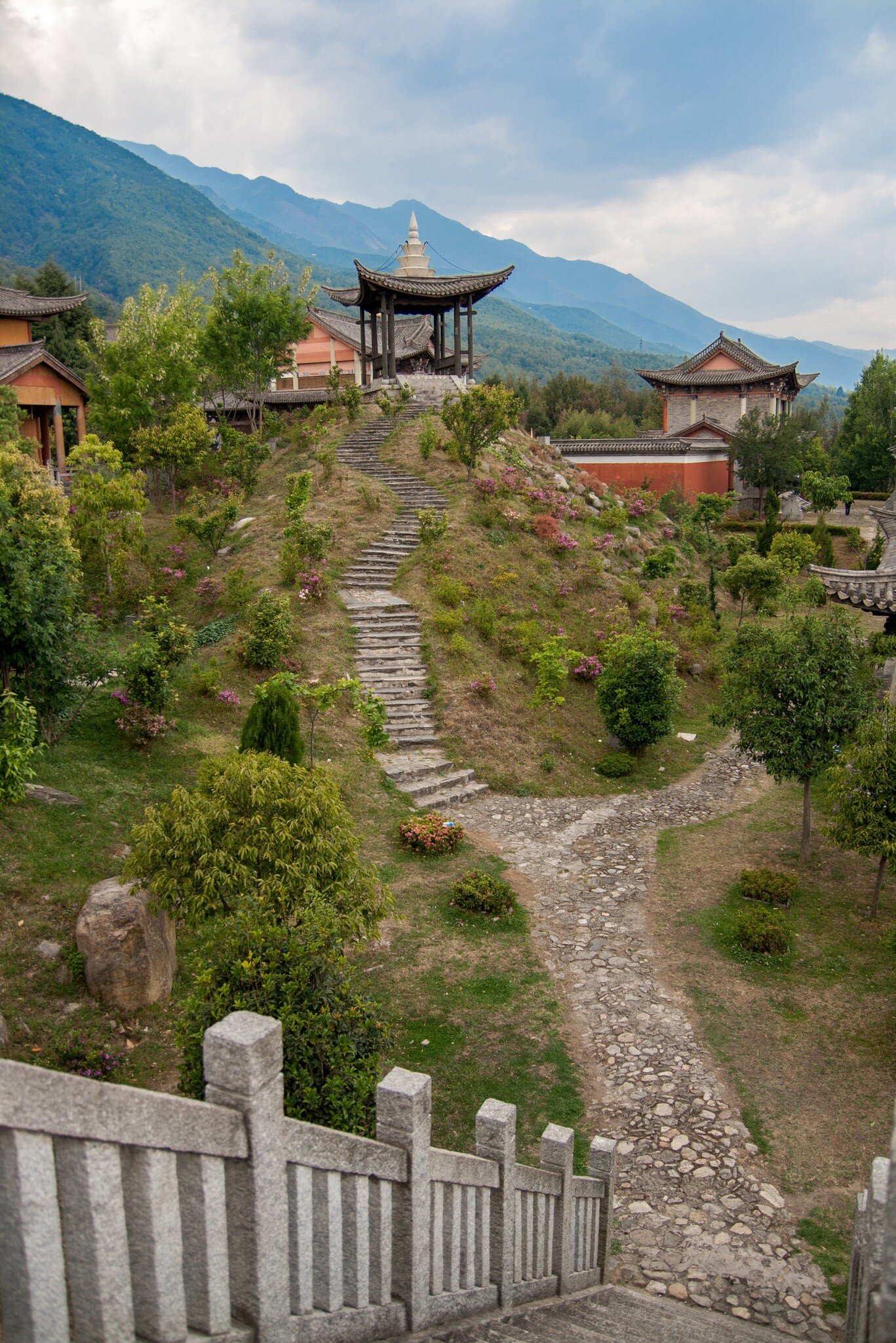 A stone path and steps lead up a flowering hillside to a pagoda-roofed pavilion, with green mountains behind.