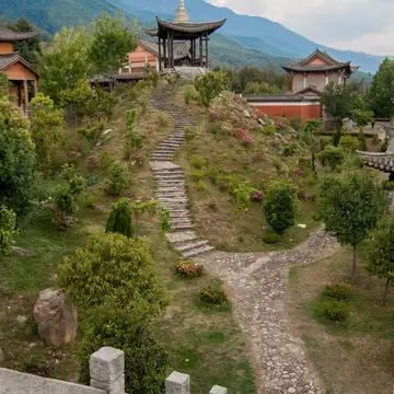 A stone path and steps lead up a flowering hillside to a pagoda-roofed pavilion, with green mountains behind.