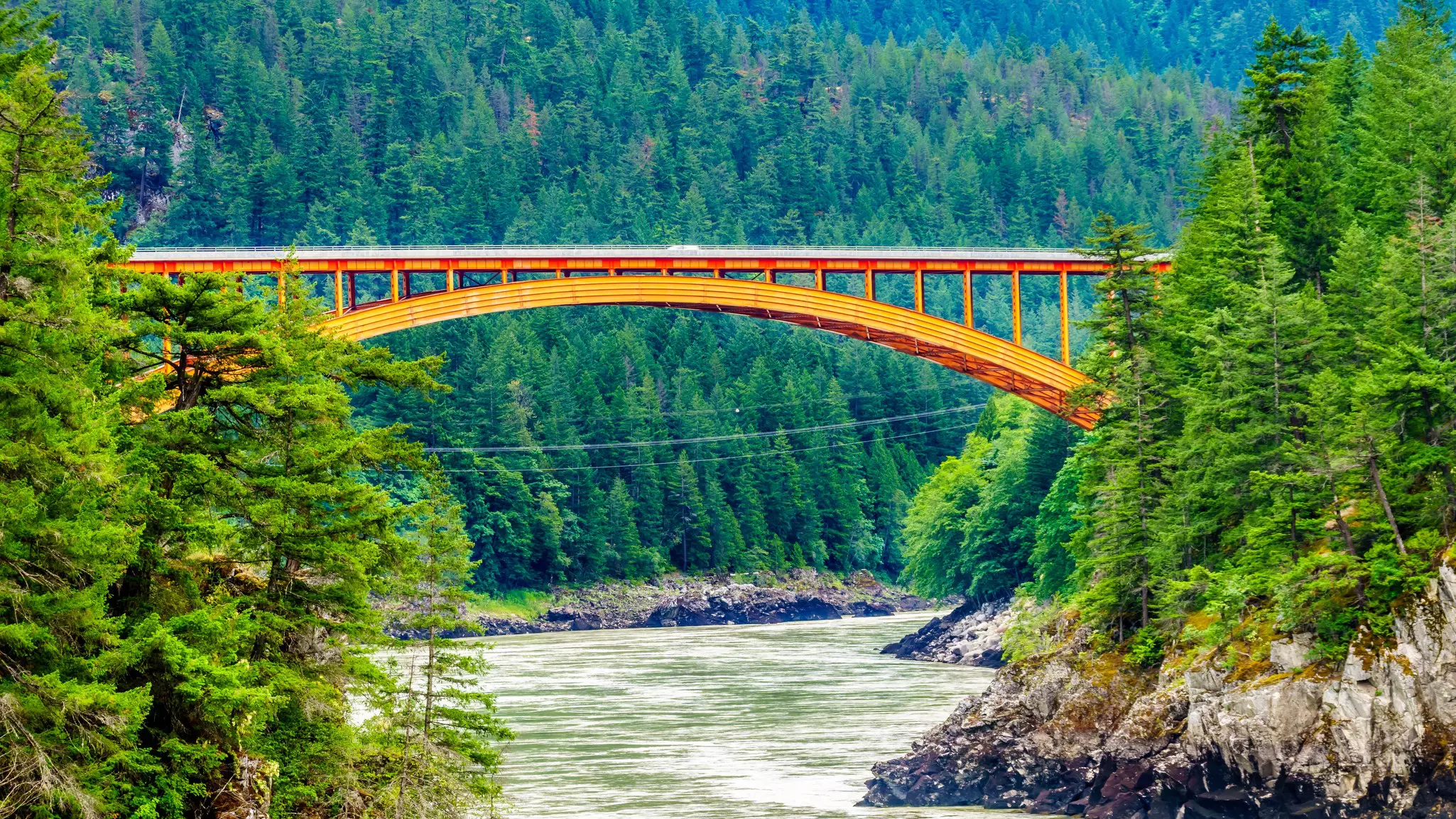 A reddish, arched steel structure of a bridge over a river with rocks and evergreens on the banks and in the distance on an overcast day.