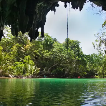 View from inside a cave mouth, with hanging rock formations above calm green water and dense tropical forest beyond.