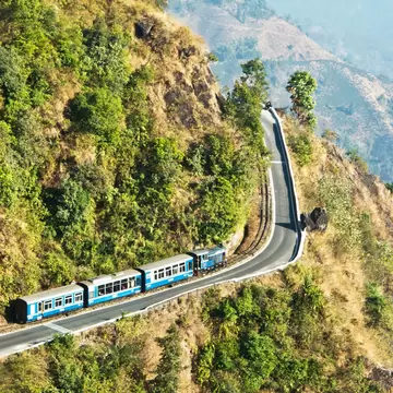 A blue train with four railcars curves around a green hill above a valley.