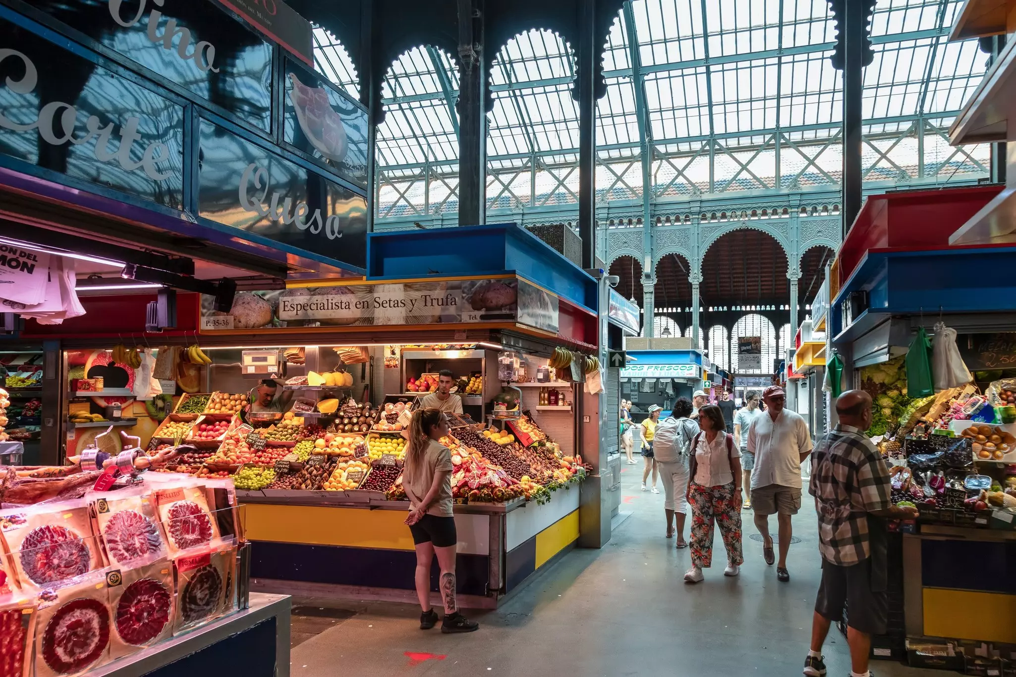 People walk by stalls in a covered market. Elaborate columns and skylights are seen in the background.