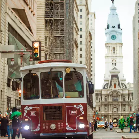 Vintage bus with red upper and red lower driving towards the camera with a colonial building in the background
