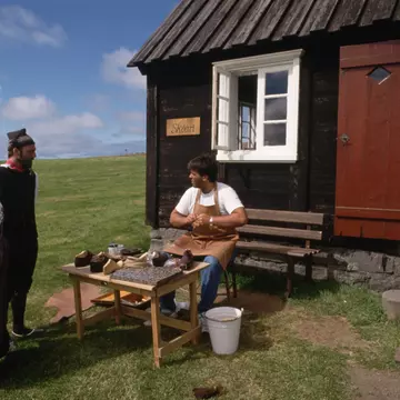 Wearing traditional dress, actors at Arbaejarsafn, or Open Air Museum, talk to a fellow actor at his shoemaking post. Reykjavik, Iceland