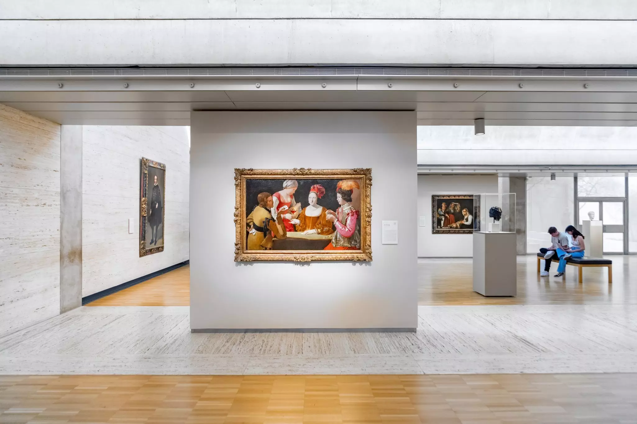 A painting depicting a card game hangs in the light-filled gallery of an art museum.
