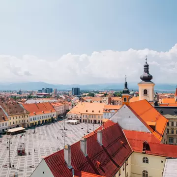 Red-tiled rooftops surround the broad Piata Mare square in Sibiu, Romania, with a baroque church tower and hills beyond.