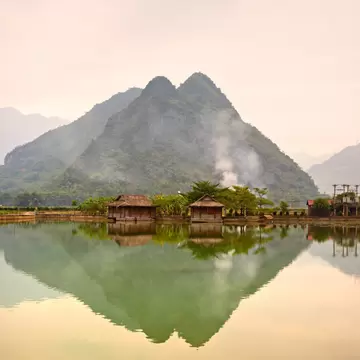 Two thatched wooden houses at the edge of a calm pond in Mai Chau, a double-peaked mountain mirrored in the water under haze.