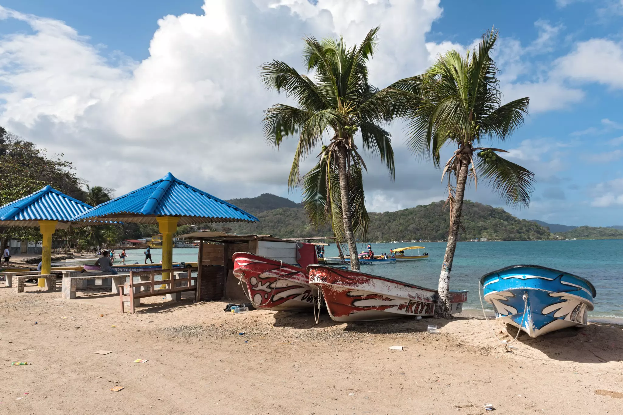 Boats pulled onto a beach between palm trees near picnic tables under blue umbrellas; the blue sea is in the background and green hills are on the far shore.
