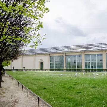 Exterior of the Musée de l’Orangerie (Oragnery Museum) in the Tuileries Gardens in Paris, France, on a cloudy day. Green grass and trees surrounding.