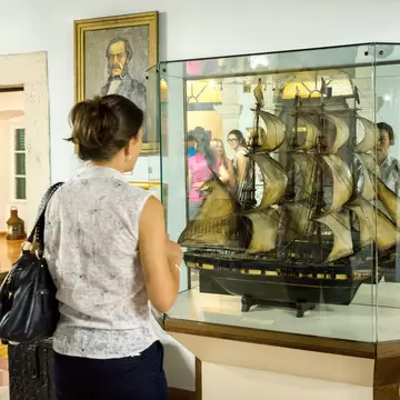 A visitor standing before a tall glass case holding a rigged model of a square-sailed warship, with a framed portrait on the wall behind.