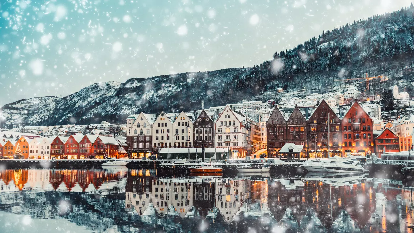 A view of the waterfront of a city on a snowy afternoon. The water reflects the warmly illuminated buildings, and a hill rises behind the built-up neighborhood.