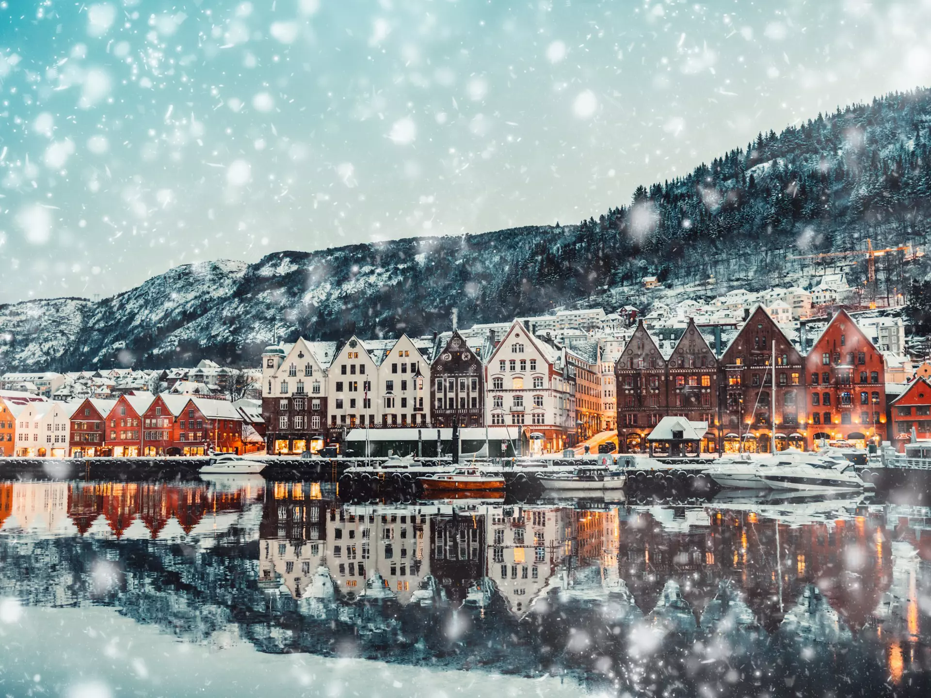 A view of the waterfront of a city on a snowy afternoon. The water reflects the warmly illuminated buildings, and a hill rises behind the built-up neighborhood.