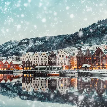 A view of the waterfront of a city on a snowy afternoon. The water reflects the warmly illuminated buildings, and a hill rises behind the built-up neighborhood.