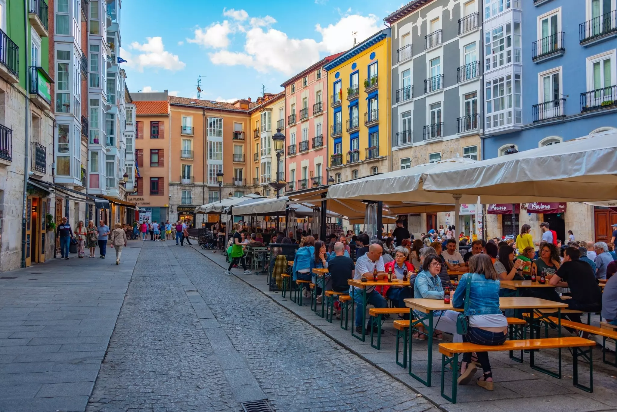 People sit at picnic tables under umbrellas in a square in Spain that has pastel-colored buildings.