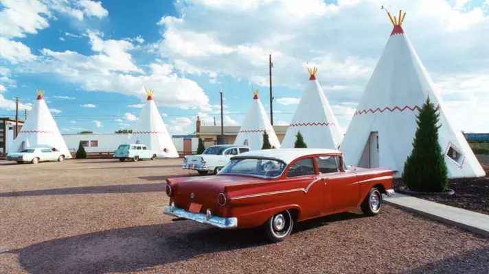 Vintage cars are parked in a lot in front of motel rooms shaped like tepees.