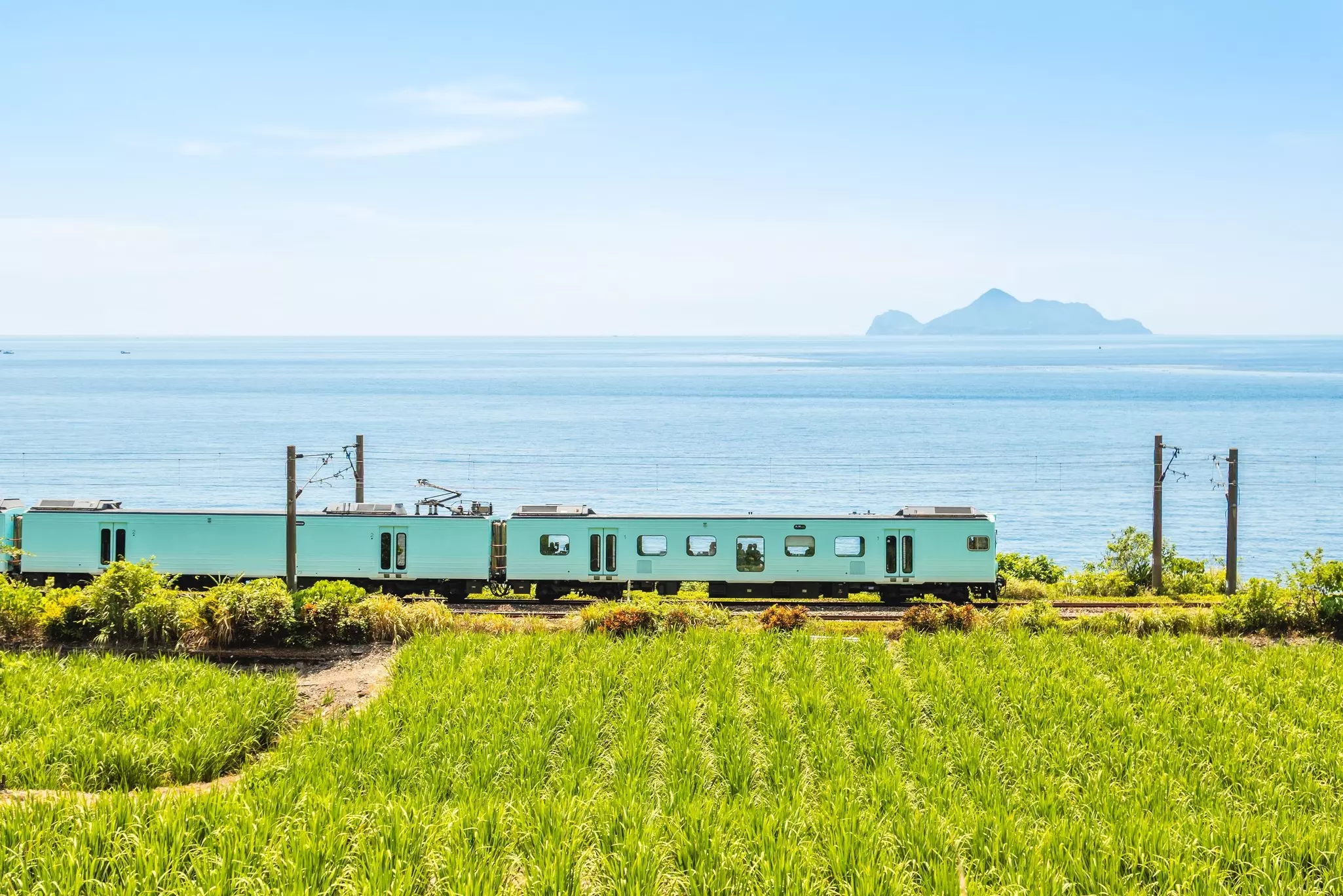A turquoise-colored train carriage runs along a track beside the sea. 