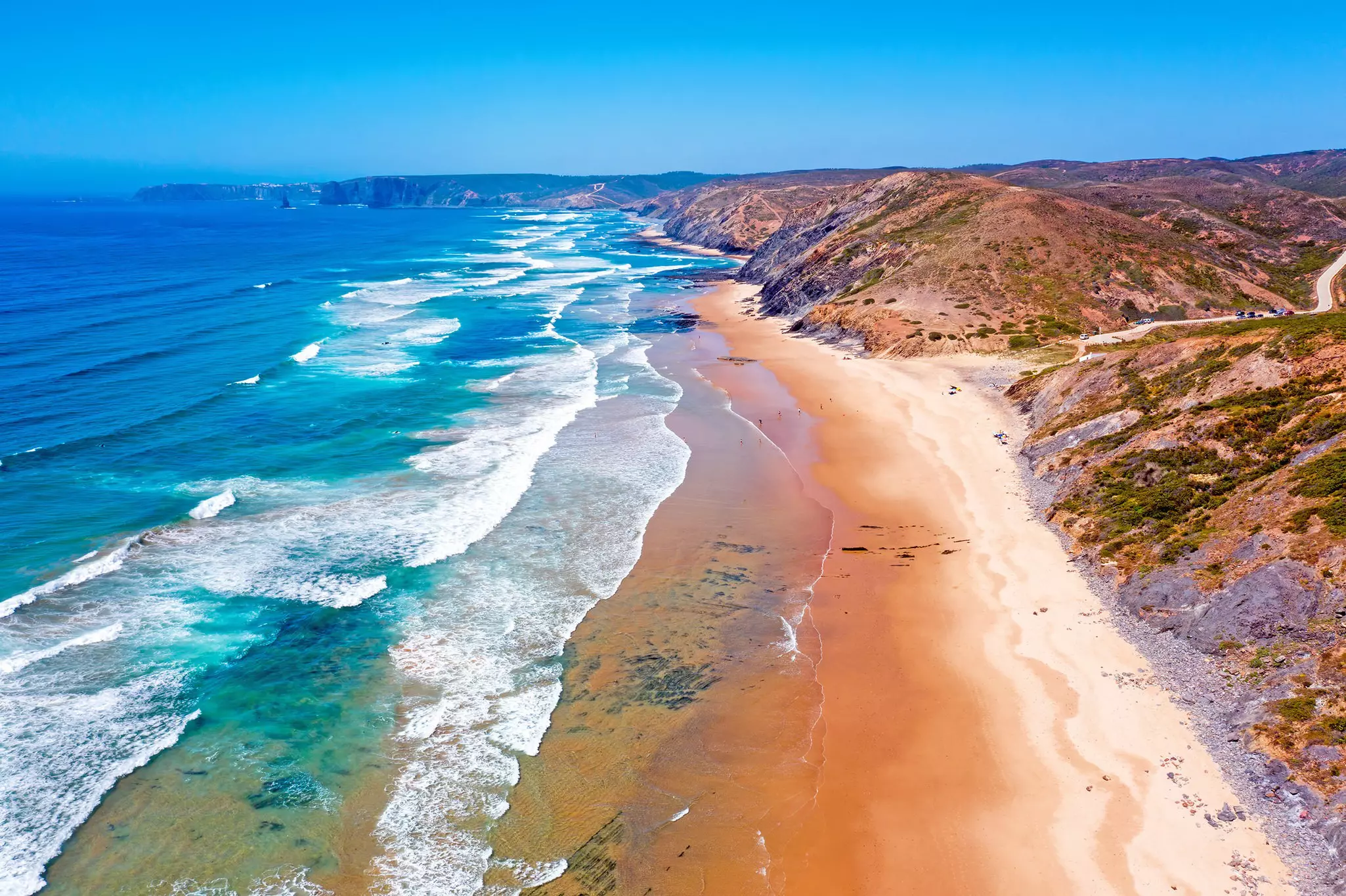 Aerial view of a beach with reddish sand, the ocean to the left, and grassy cliffs to the right.