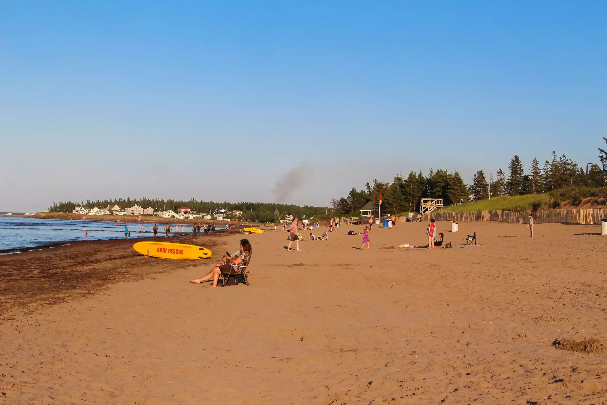 People sit on a wide beach with golden sand, with houses and businesses seen in the distance.