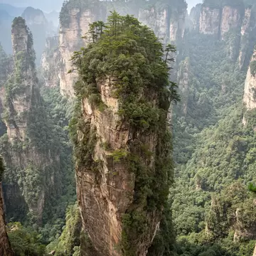A tall, sheer rock column crowned with pine trees stands in a green valley, more stone pillars fading into haze behind.
