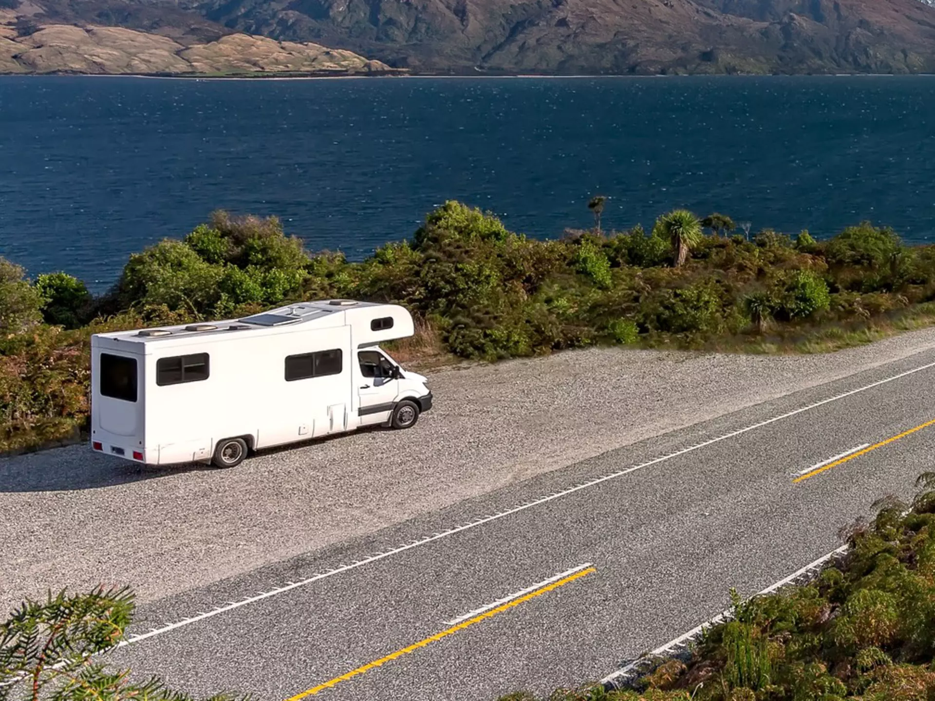 A white camper-van parked in a lay-by viewpoint on the banks of Lake Wānaka with dramatic snow capped peaks in the distance, Queenstown NZ