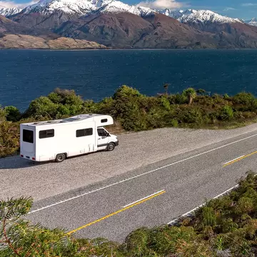 A white camper-van parked in a lay-by viewpoint on the banks of Lake Wānaka with dramatic snow capped peaks in the distance, Queenstown NZ