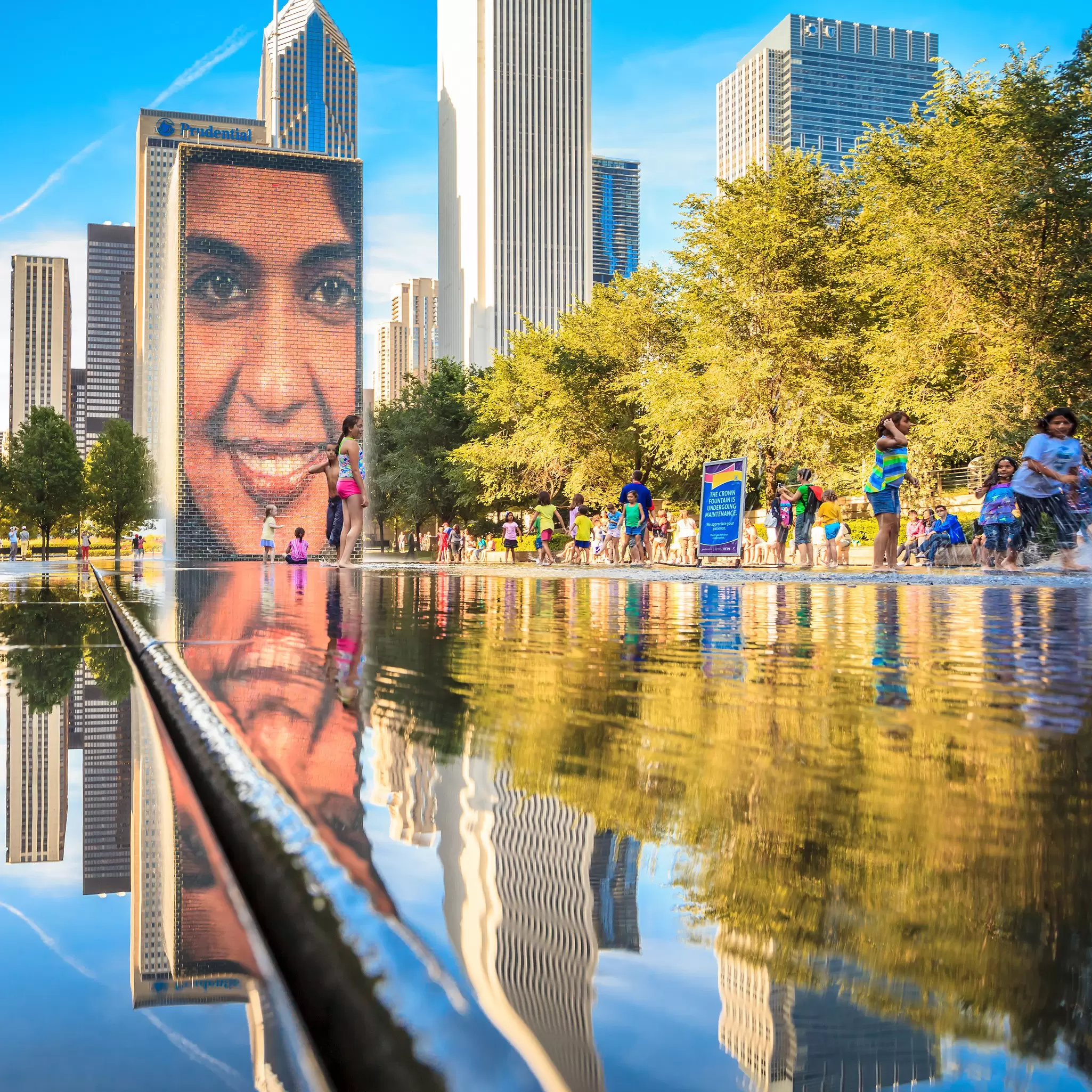 A large video screen with a smiling face stands above a water fountain in a park