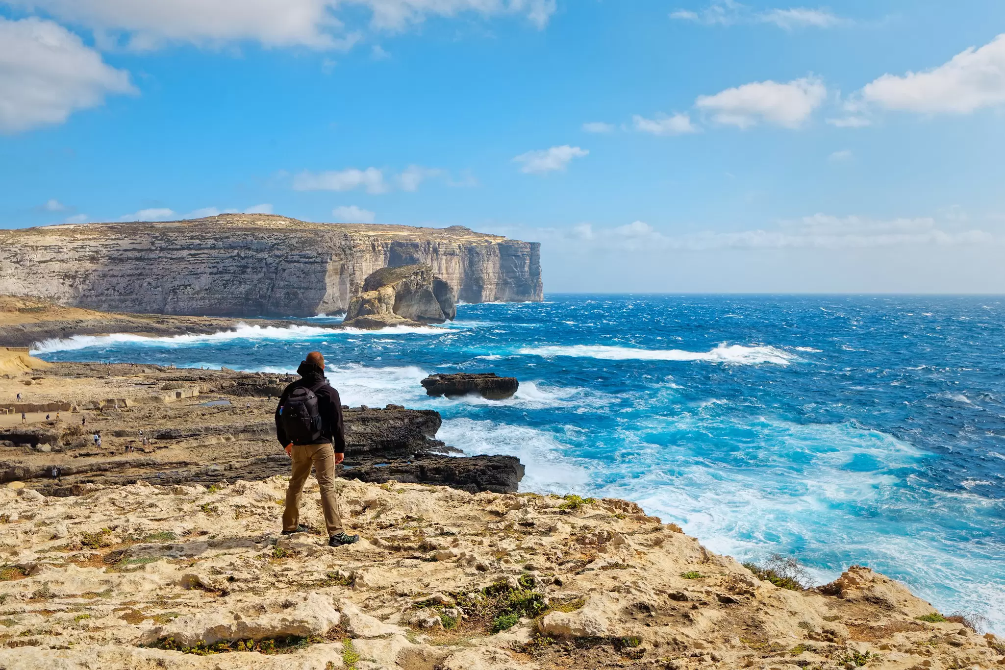 A hiker is seen from behind standing on a cliff and looking out at the rough waters in a bay below