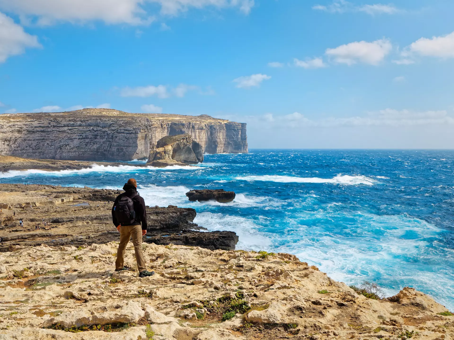 A hiker is seen from behind standing on a cliff and looking out at the rough waters in a bay below