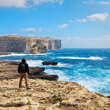 A hiker is seen from behind standing on a cliff and looking out at the rough waters in a bay below