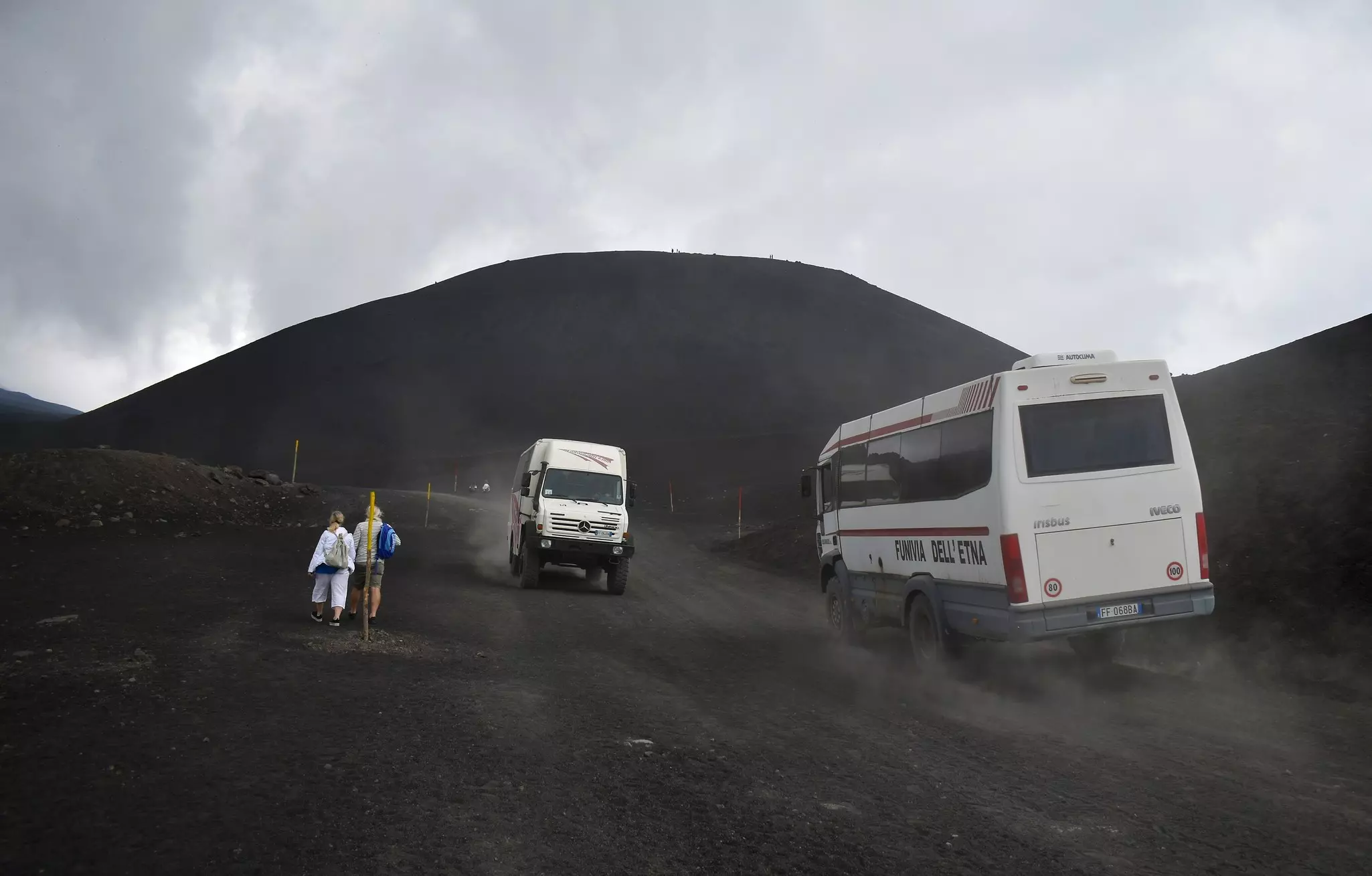 Two white off-road vehicles drive down a dirt road on the slope of a volcano. Two people walk to the side of the road.