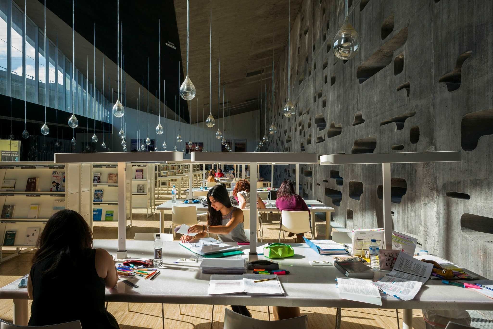 Patrons at tables in a library with teardrop-like mirrors hanging from its ceiling and a concrete wall with holes carved in it