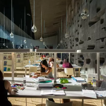 Patrons at tables in a library with teardrop-like mirrors hanging from its ceiling and a concrete wall with holes carved in it