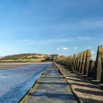Concrete WWII anti-boat pylons beside a causeway across wet sand at low tide, leading to wooded Cramond Island near Edinburgh
