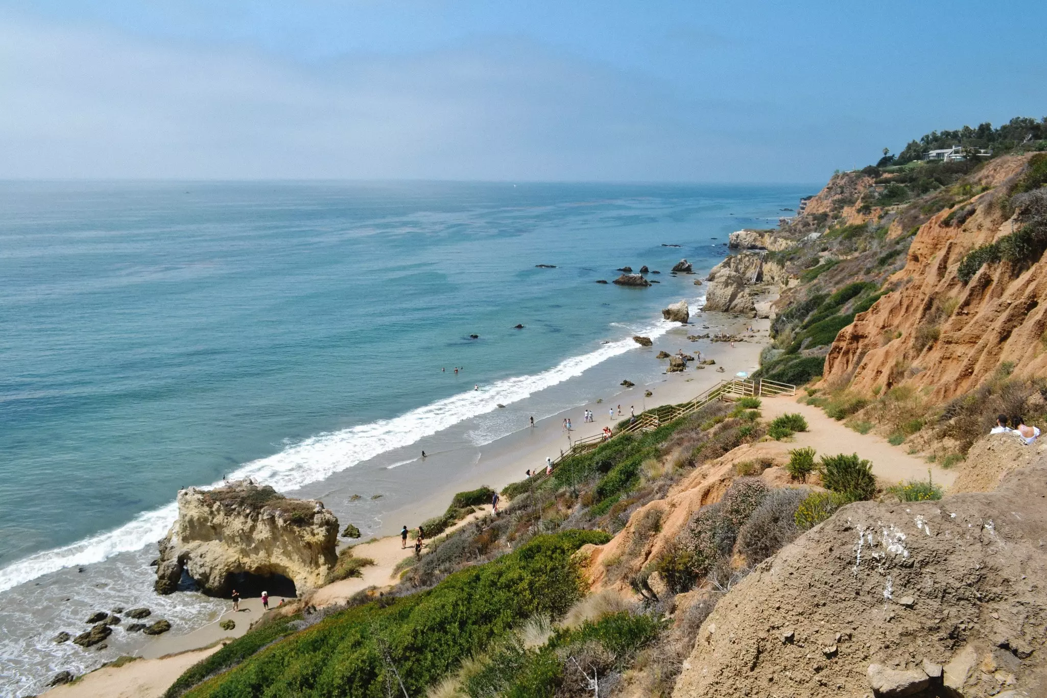 Aerial view of a dirt- and brush-filled hillside with steep steps in the distance leading to a coved, sandy beach and the ocean beyhond.