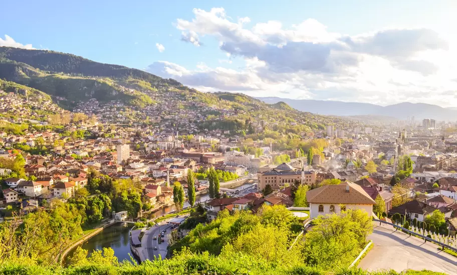 Sarajevo seen from a hillside, red-roofed houses climbing green slopes, Miljacka River and city centre below