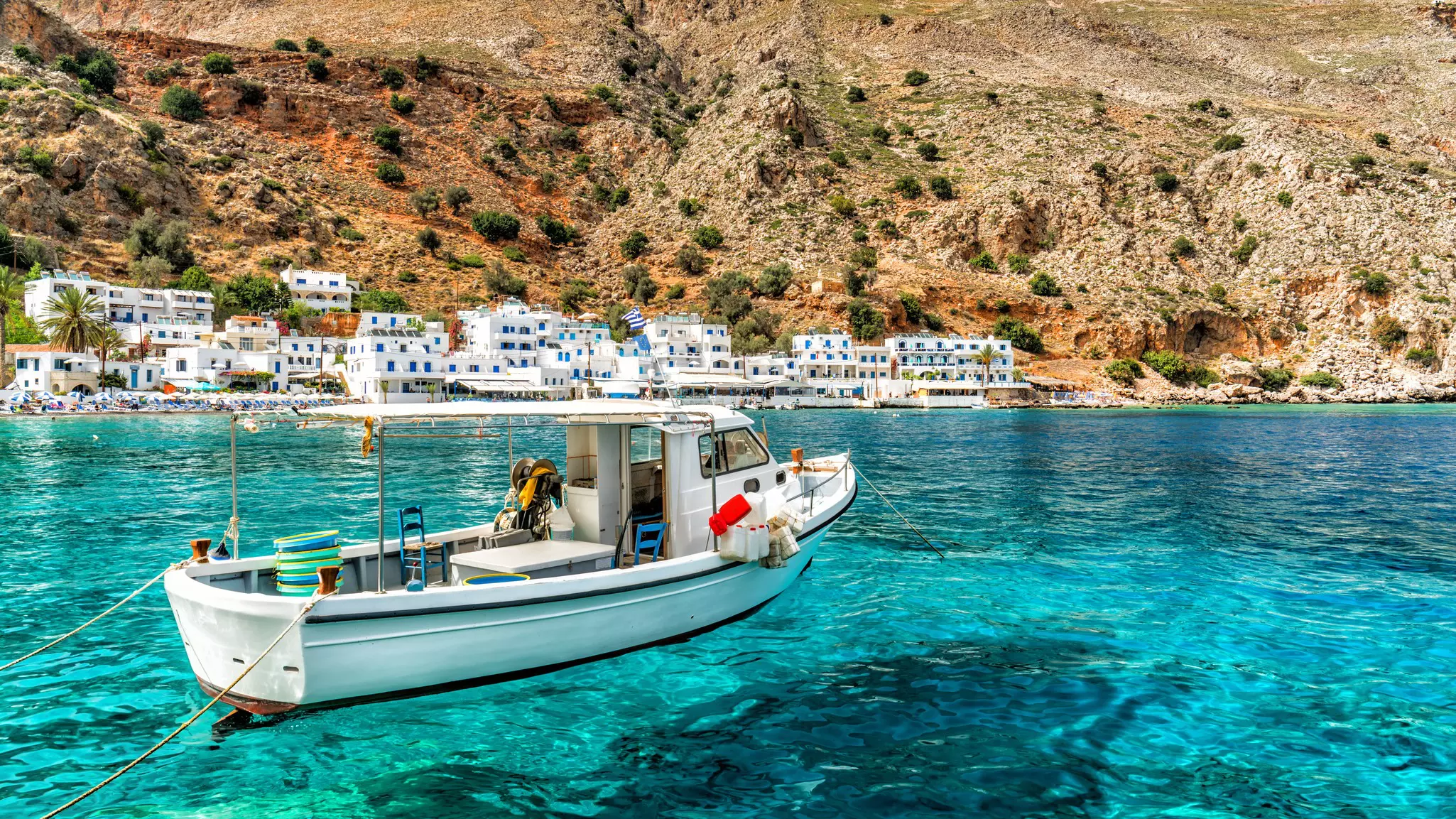 A white fishing boat is moored in shallow, clear waters by a small village with white waterfront houses.