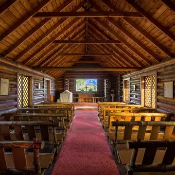 Log interior of Chapel of the Transfiguration, pews leading to an altar window with a cross and Teton peaks beyond
