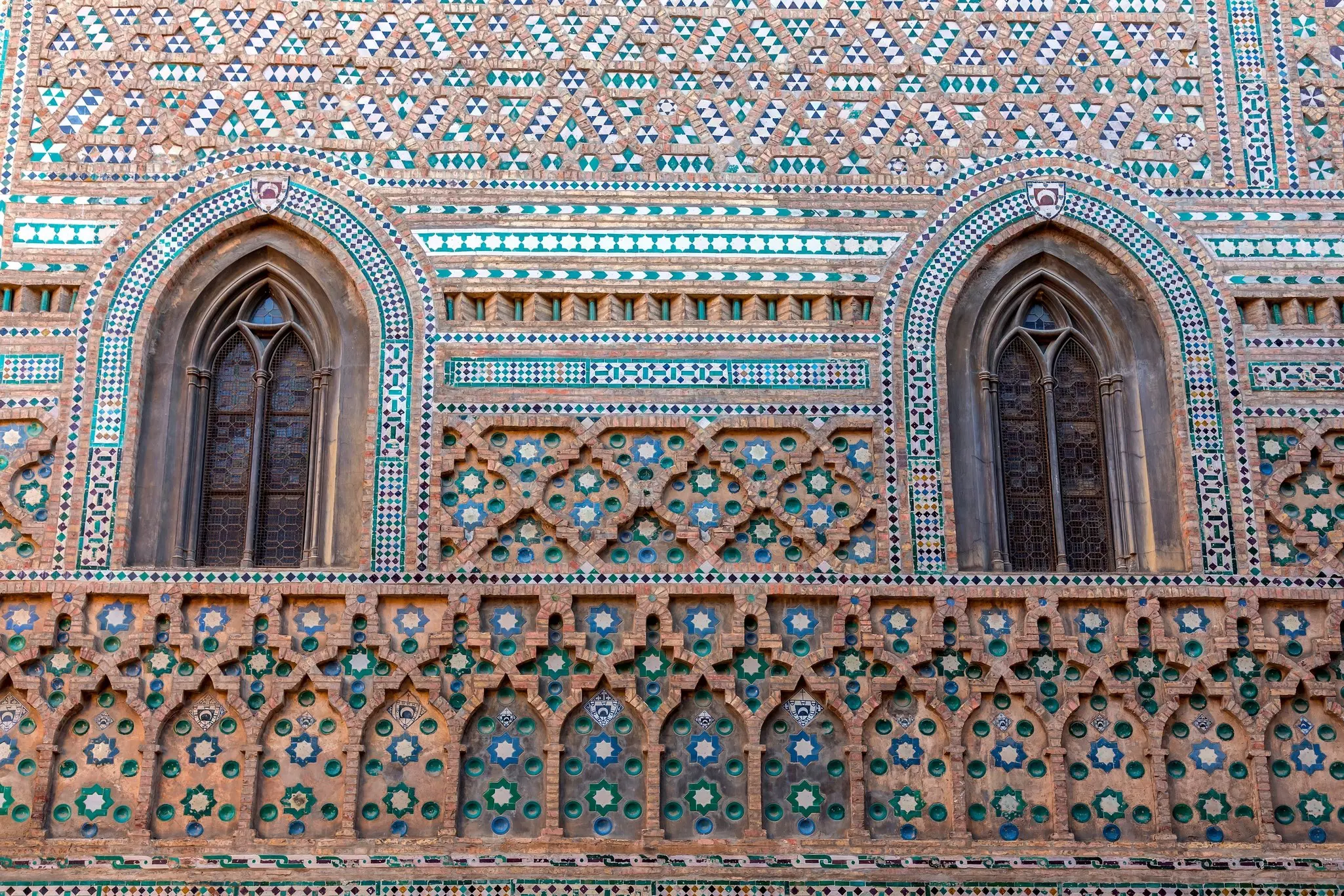 Exterior wall of a cathedral building with classic brickwork and colourful ceramics in complex geometric patterns around two narrow windows.