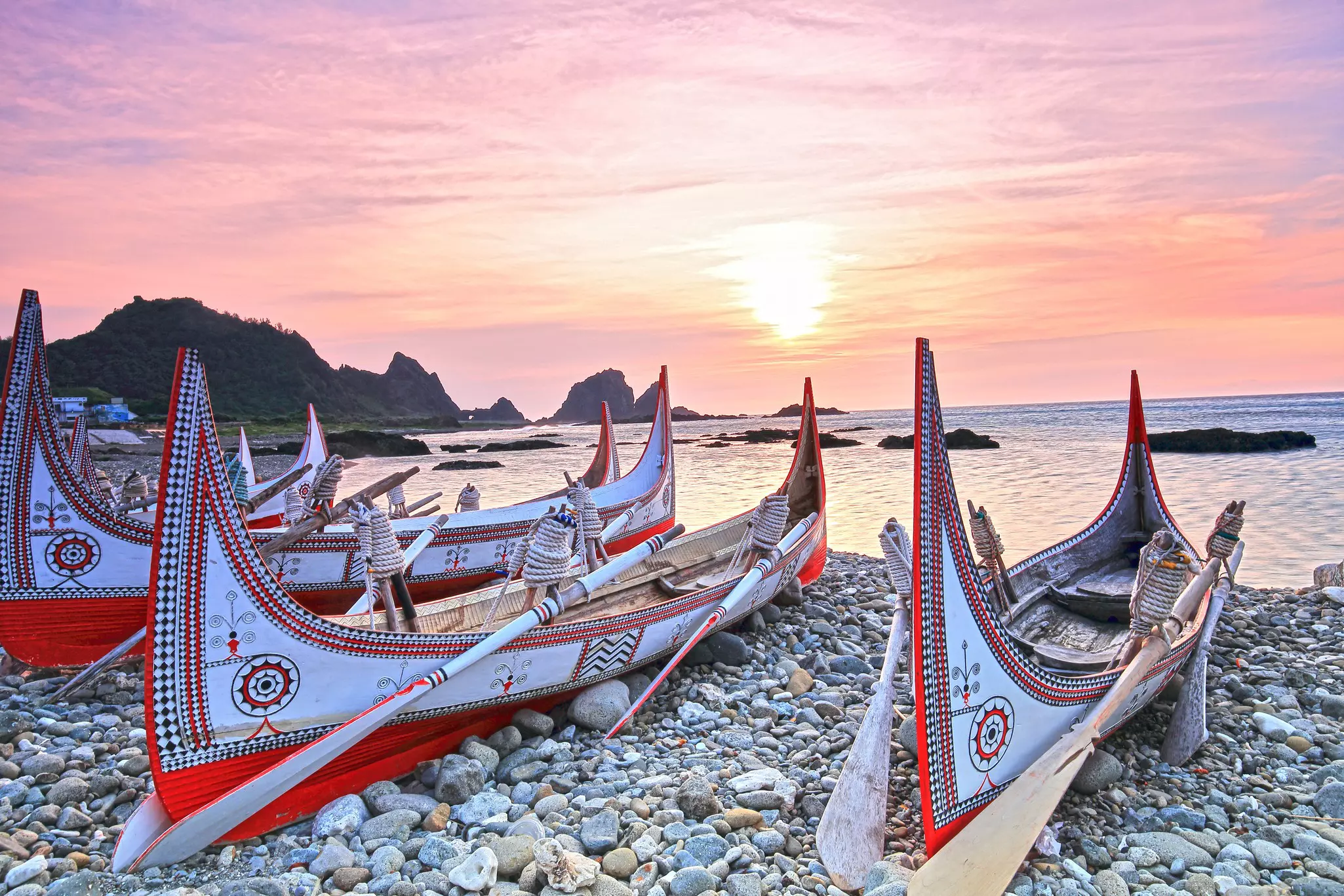 Canoes painted with traditional designed sit on a rocky beach on an island. The sunset is visible in the distance.