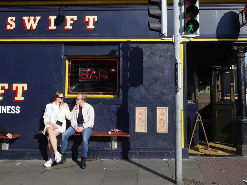 Two people sit on a bench outside a navy blue bar on a sunny day.