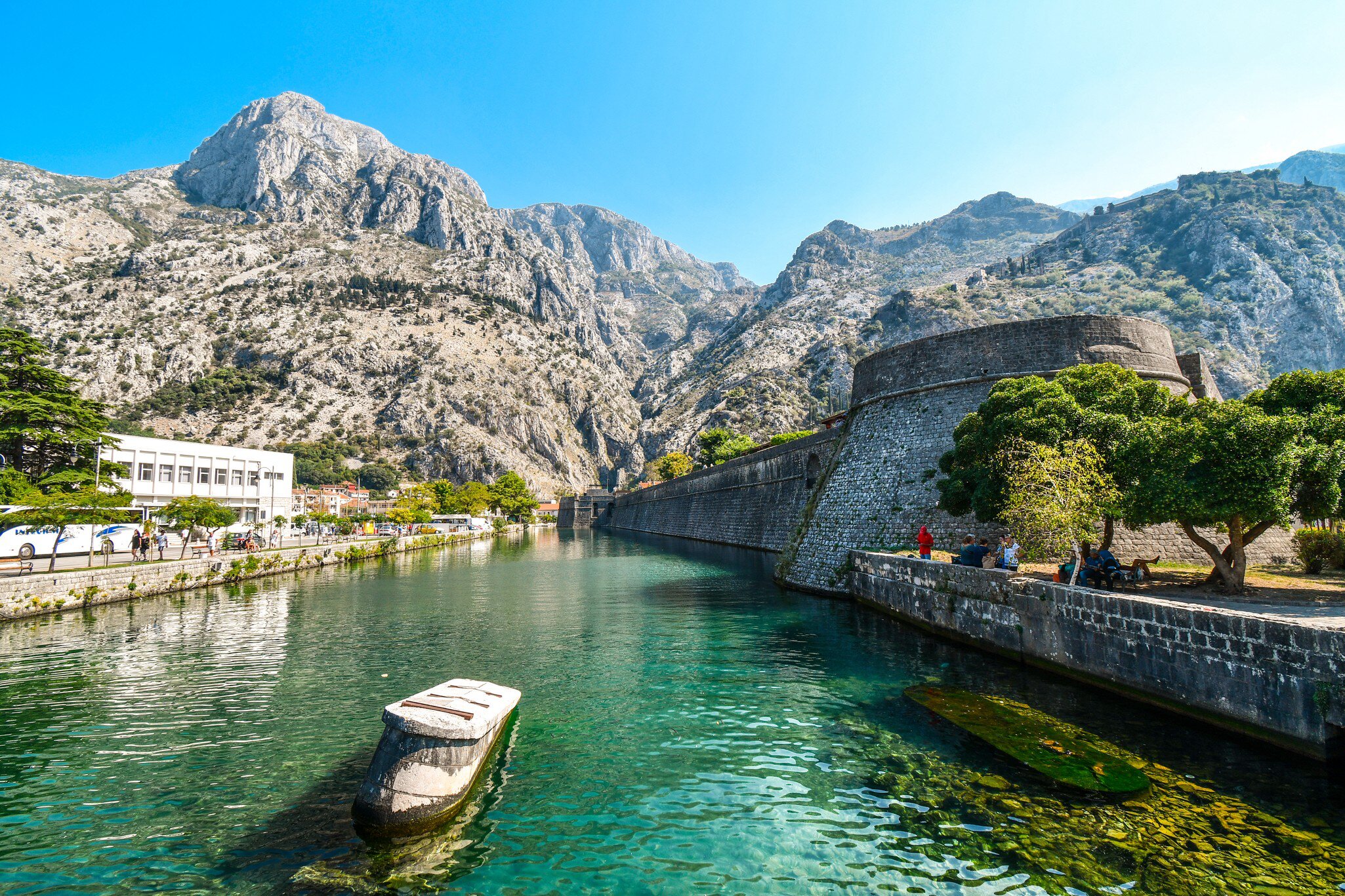 A stone bastion and long curtain wall rising from clear green water, with bare grey mountains behind and people sitting on the far bank.