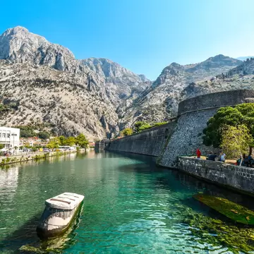 A stone bastion and long curtain wall rising from clear green water, with bare grey mountains behind and people sitting on the far bank.
