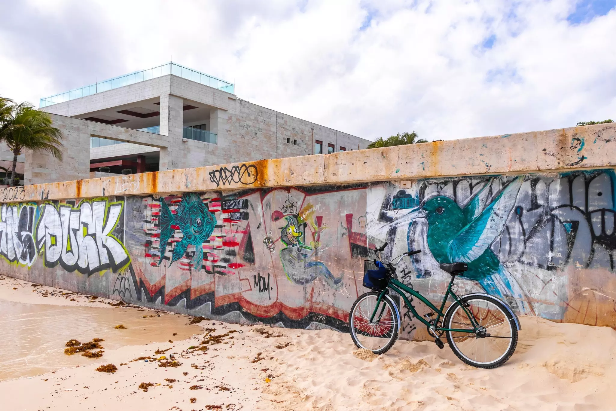 A bicycle is parked on sand, leaning against a wall covered in bright graffiti.