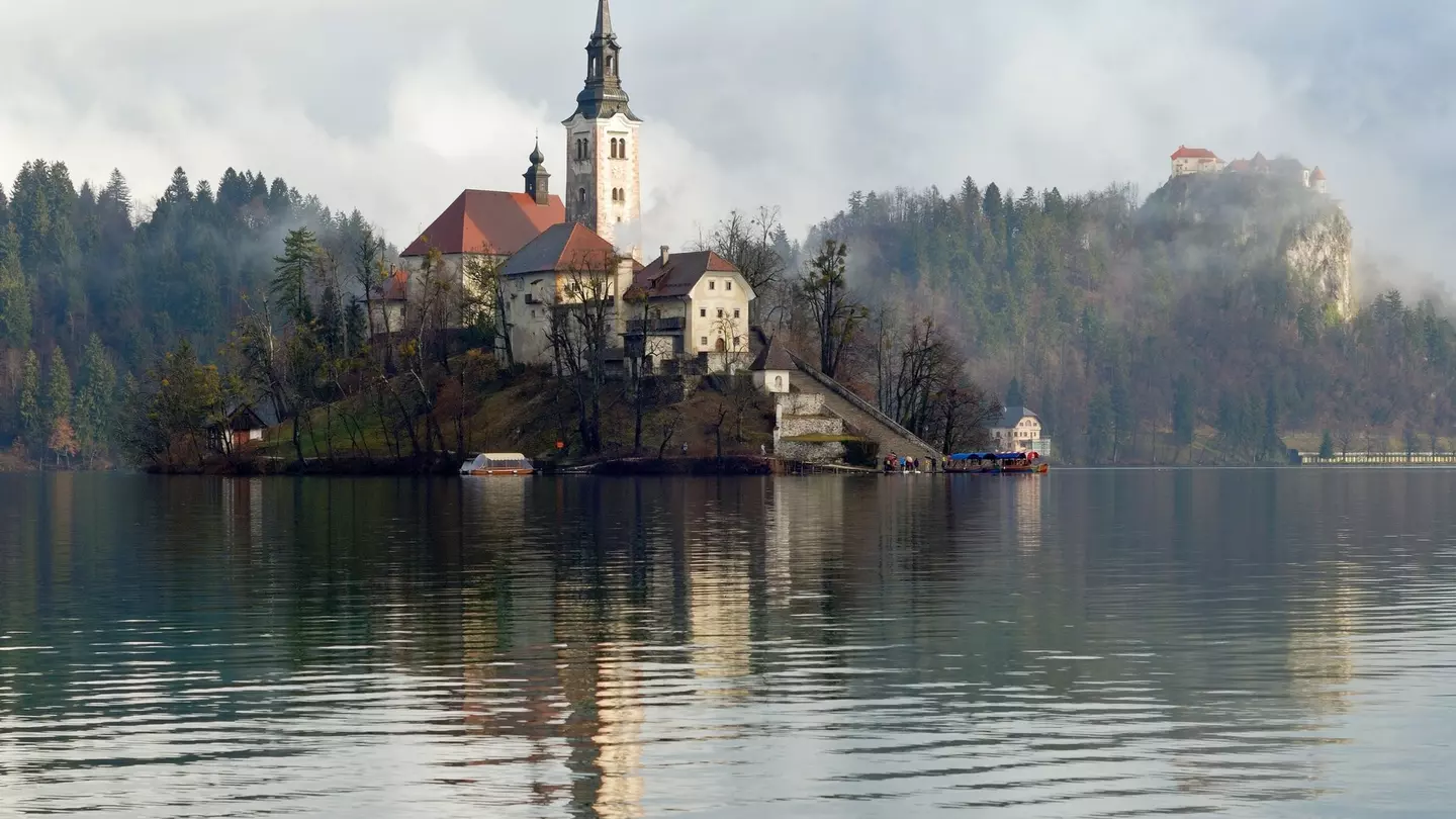 A church located on an island in the middle of a lake with the castle standing on a clifftop nearby shrouded in mist.