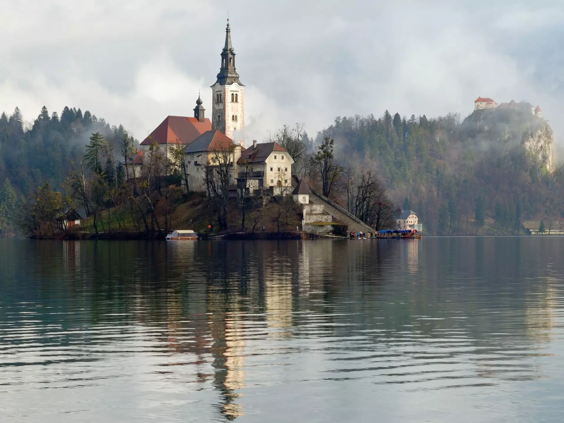 A church located on an island in the middle of a lake with the castle standing on a clifftop nearby shrouded in mist.