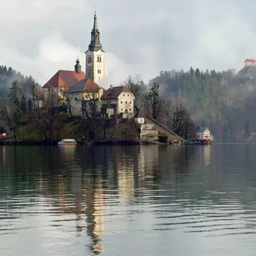 A church located on an island in the middle of a lake with the castle standing on a clifftop nearby shrouded in mist.